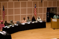 [ai] A panel of speakers sitting at a long table in a conference setting, with American flags in the background. The speakers are diverse in gender and attire, discussing topics with documents and notes in front of them.